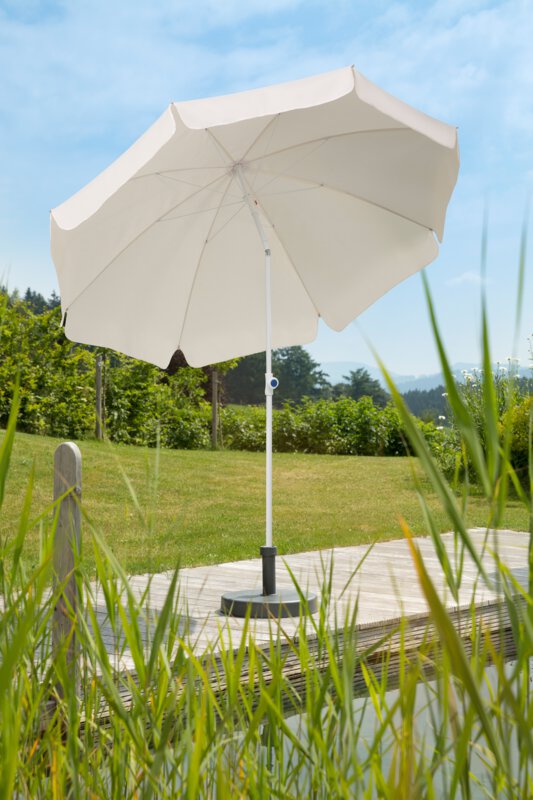 Schneider Ibiza parasol in ecru, 240 cm in diameter, on a wooden terrace by a natural swimming pool. Reeds in the foreground, meadow and various green plants in the background.