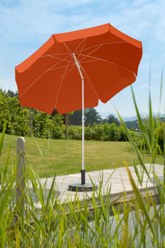 Schneider Ibiza parasol in terracotta, 240 cm in diameter, on a wooden terrace by a natural swimming pool. Reeds in the foreground, meadow and various green plants in the background.