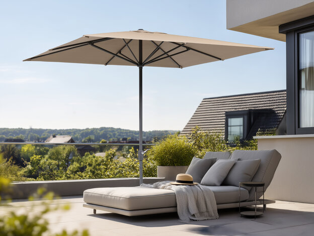 On a terrace, the Schneider Korsika parasol provides shade over a comfortable lounge chair with a hat and a blanket. Houses and trees are visible in the background.