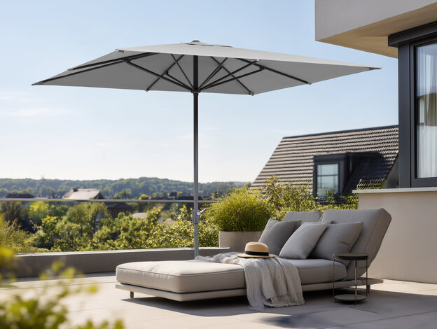 On a terrace, the Schneider Korsika parasol provides shade over a comfortable lounge chair with a hat and a blanket. Houses and trees are visible in the background.
