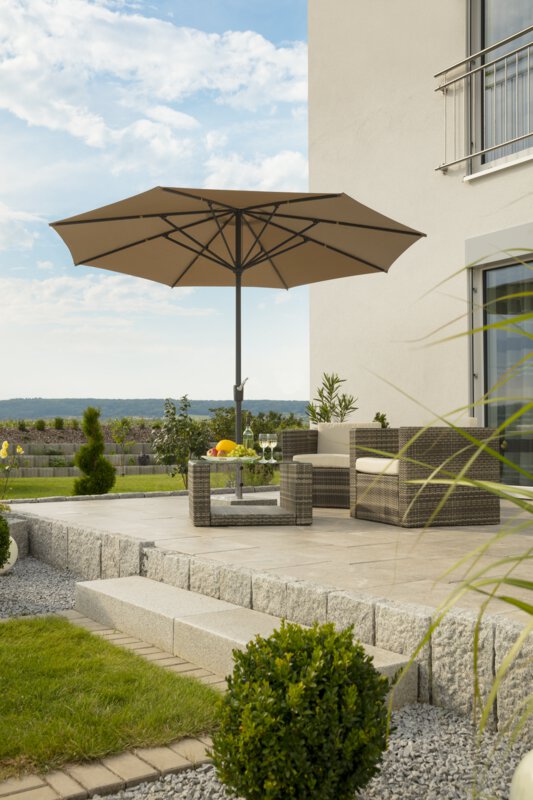 Schneider Korsika parasol in ecru, 320 cm in diameter, on a terrace with lounge furniture underneath. A fruit bowl, a bottle and glasses are on the table. A stepped retaining wall made of concrete blocks is in the background. The terrace is surrounded by grass and shrubs.