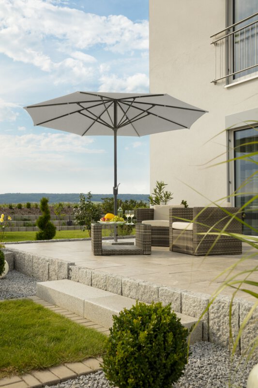 Schneider parasol Korsika in silver grey, 320 cm in diameter, on a terrace with lounge furniture below. On the table is a fruit bowl, a bottle and glasses. In the background, a stepped retaining wall made of concrete blocks. The terrace is surrounded by grass and shrubs.