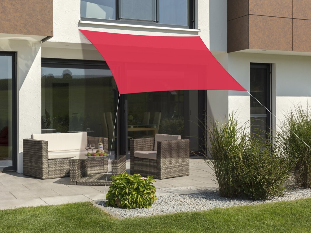Schneider Lanzarote sun sail in red, 300x250 cm, stretched over a terrace with lounge furniture. A fruit bowl with a bottle and glasses sits on the table. Small shrubs and a meadow are in the foreground.