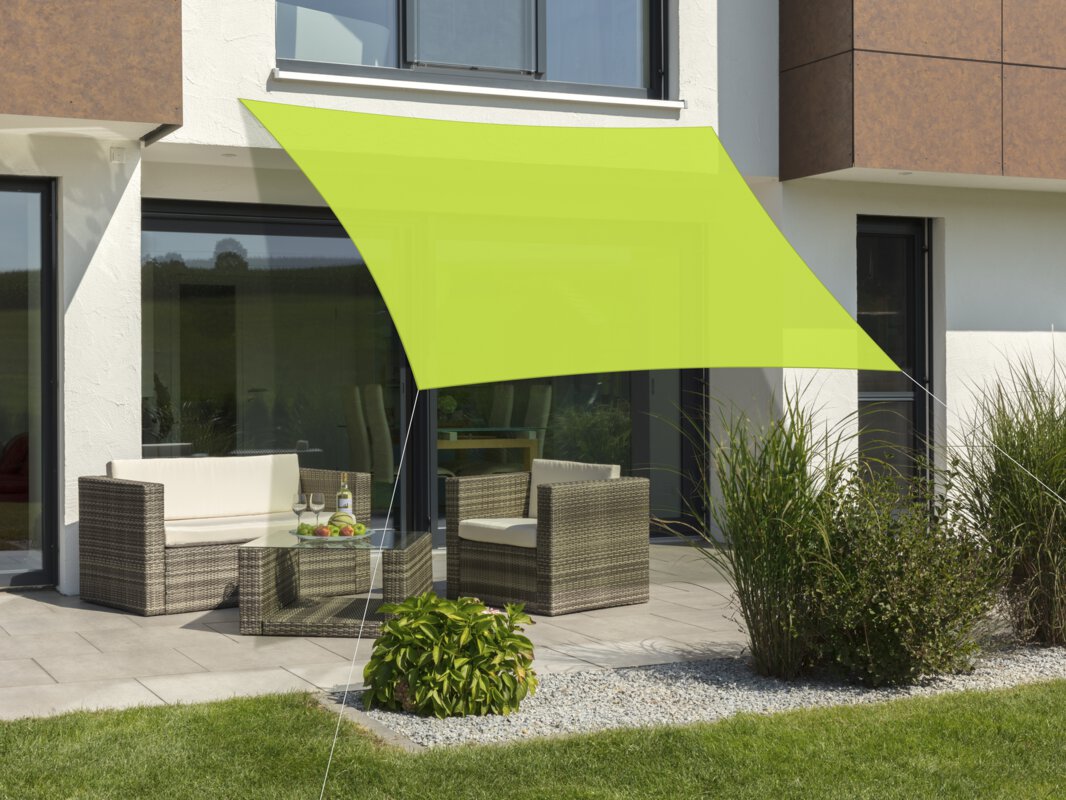 Schneider Lanzarote sun sail in apple green, 360x360 cm, stretched over a terrace with lounge furniture. A fruit bowl with a bottle and glasses sits on the table. Small shrubs and a meadow are in the foreground.