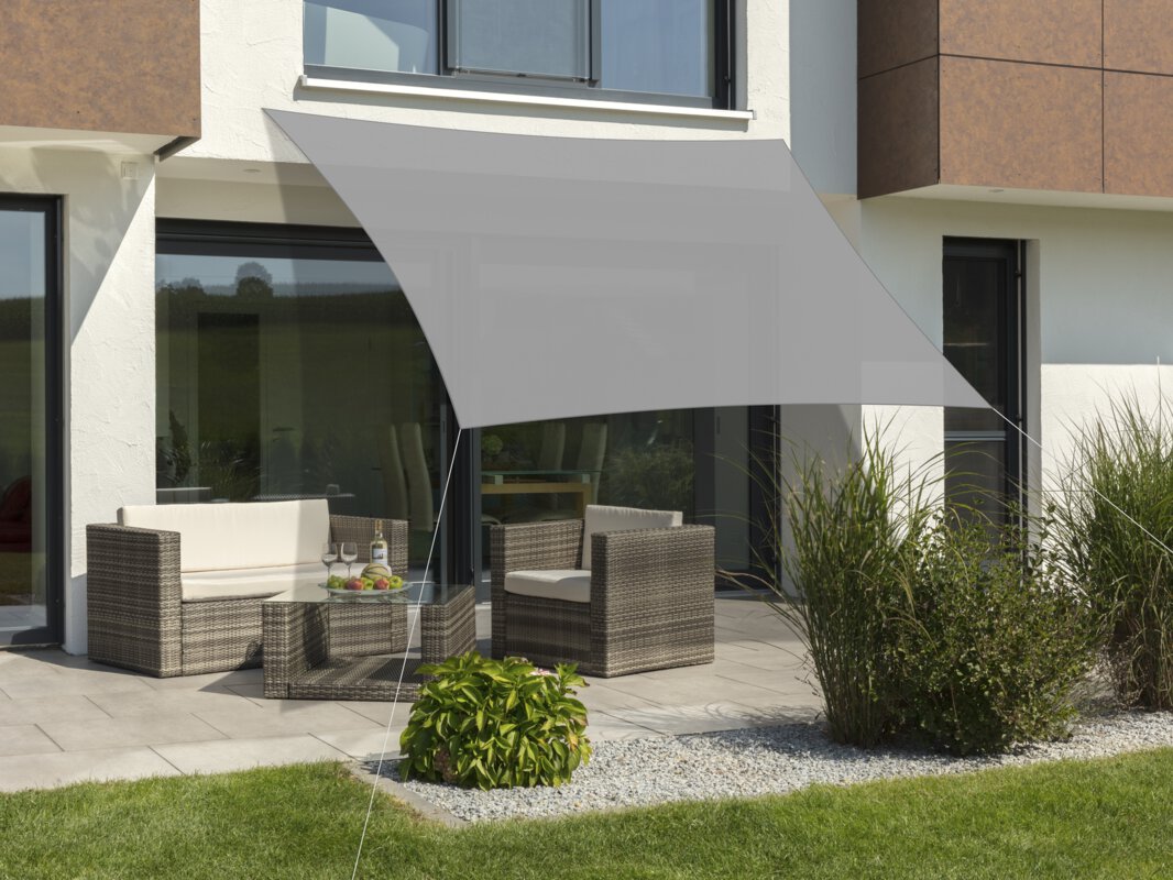 Schneider Lanzarote sun sail in silver grey, 360x360 cm, stretched over a terrace with lounge furniture. A fruit bowl with a bottle and glasses sits on the table. Small shrubs and a meadow are in the foreground.