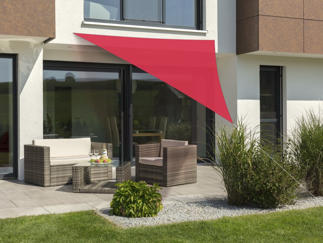Schneider Sonnensegel Lanzarote in Rot, 360x360x360 cm, gespannt über eine Terrasse mit Lounge-Möbeln. Auf dem Tisch befindet sich eine Obstschale mit einer Flasche und Gläsern. Im Vordergrund kleine Sträucher und Wiese.