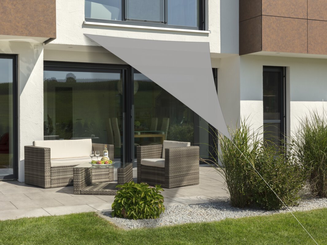 Schneider Lanzarote sun sail in silver grey, 400x400x300 cm, stretched over a terrace with lounge furniture. A fruit bowl with a bottle and glasses sits on the table. Small shrubs and a meadow are in the foreground.