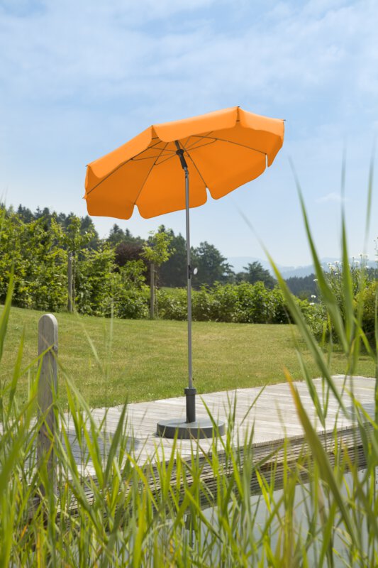 Schneider Sonnenschirm Locarno in Mandarine, 150 cm Durchmesser, auf einer Holzterrasse an einem Naturpool. Im Vordergrund Schilf, im Hintergrund Wiese und verschiede Grünpflanzen.