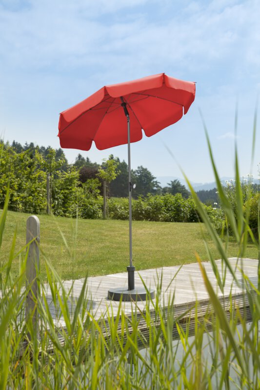 Schneider Sonnenschirm Locarno in Rot, 150 cm Durchmesser, auf einer Holzterrasse an einem Naturpool. Im Vordergrund Schilf, im Hintergrund Wiese und verschiede Grünpflanzen.