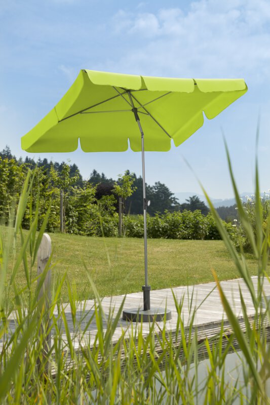 Schneider Locarno parasol in apple green, 180x120 cm, on a wooden terrace by a natural swimming pool. Reeds in the foreground, meadow and various green plants in the background.