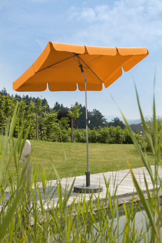 Schneider Sonnenschirm Locarno in Mandarine, 180x120 cm, auf einer Holzterrasse an einem Naturpool. Im Vordergrund Schilf, im Hintergrund Wiese und verschiede Grünpflanzen.