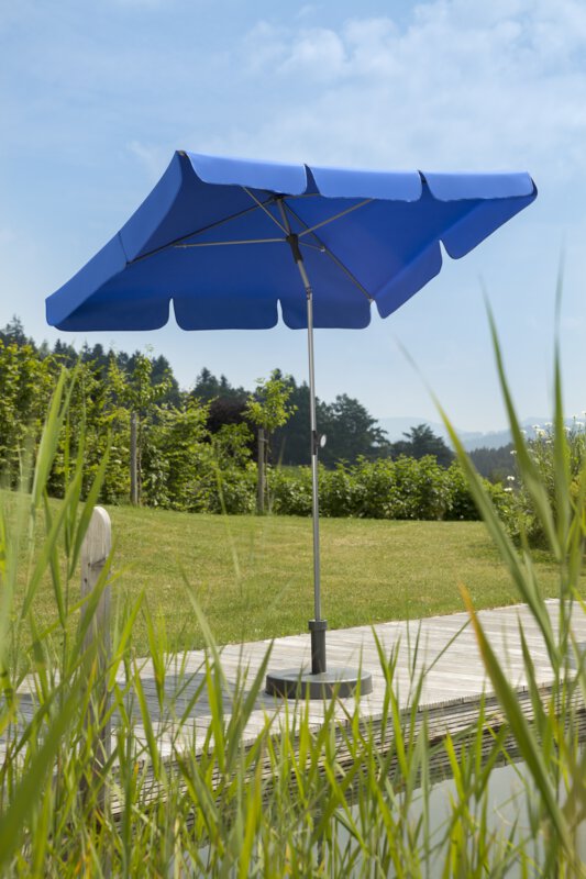 Schneider Locarno parasol in royal blue, 180x120 cm, on a wooden terrace by a natural swimming pool. Reeds in the foreground, meadow and various green plants in the background.