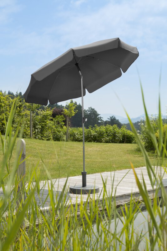 Schneider Locarno parasol in anthracite, 200 cm in diameter, on a wooden terrace by a natural swimming pool. Reeds in the foreground, meadow and various green plants in the background.