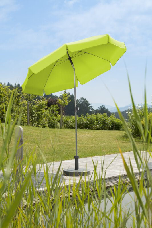 Schneider Locarno parasol in apple green, 200 cm in diameter, on a wooden terrace by a natural swimming pool. Reeds in the foreground, meadow and various green plants in the background.
