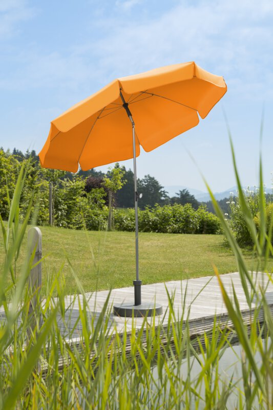 Schneider Locarno parasol in mandarin, 200 cm in diameter, on a wooden terrace by a natural swimming pool. Reeds in the foreground, meadow and various green plants in the background.