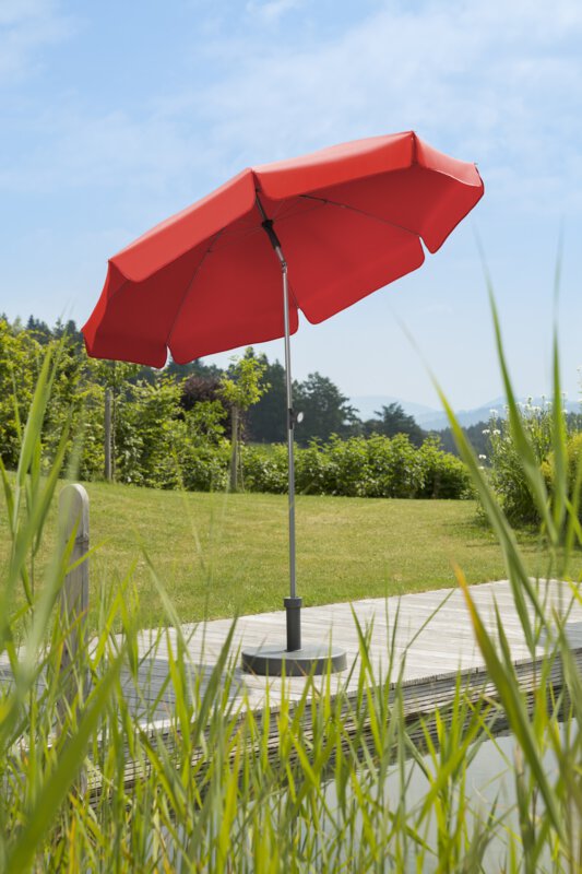 Schneider Locarno parasol in red, 200 cm in diameter, on a wooden terrace by a natural swimming pool. Reeds in the foreground, meadow and various green plants in the background.