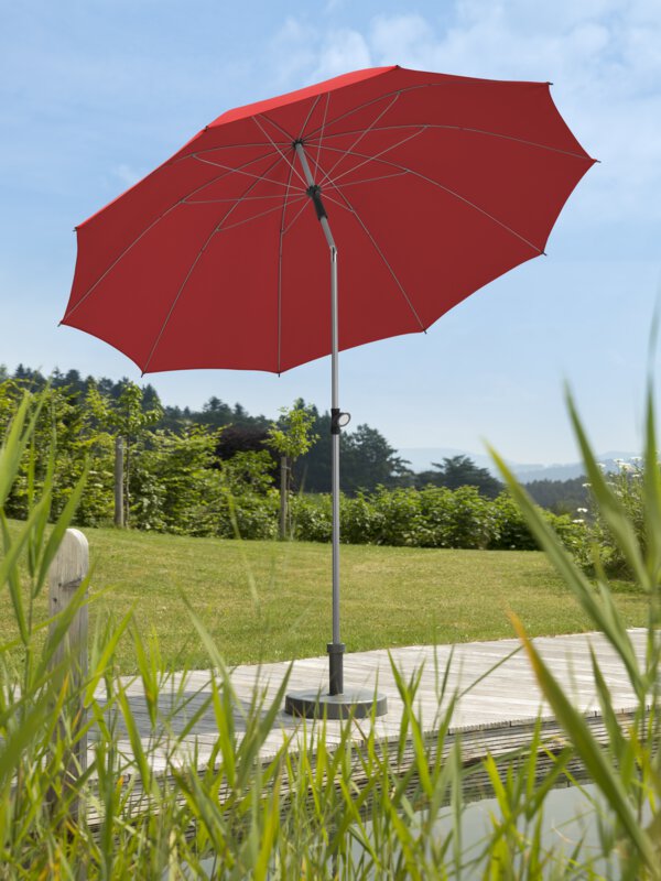 Schneider parasol Locarno in red, 220 cm in diameter, on a wooden terrace by a natural swimming pool. Reeds in the foreground, meadow and various green plants in the background.