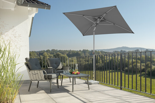 Schneider Sonnenschirm Lumi in Anthrazit, 210x130 cm, auf einer Dachterrasse. Im Schatten des Schirms stehen zwei Stühle mit grauen Sitzpolstern und ein kleiner Tisch mit Obstschale, Karaffe und Gläsern. Im Hintergrund Wiese, Wald und ein Hügel unter strahlend blauem Himmel.