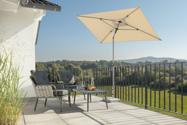 Schneider Sonnenschirm Lumi in Natur, 210x130 cm, auf einer Dachterrasse. Im Schatten des Schirms stehen zwei Stühle mit grauen Sitzpolstern und ein kleiner Tisch mit Obstschale, Karaffe und Gläsern. Im Hintergrund Wiese, Wald und ein Hügel unter strahlend blauem Himmel.