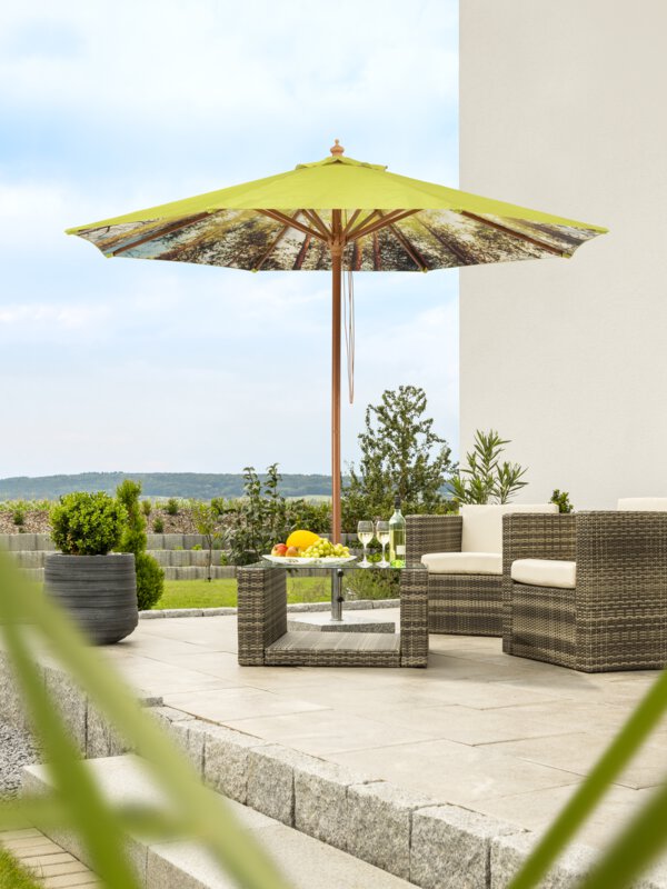A Schneider Malaga parasol in forest, 300 cm in diameter, on a terrace with lounge furniture below. A fruit bowl, a bottle and glasses are on the table. In the background, a stepped retaining wall made of concrete blocks. The terrace is surrounded by grass and shrubs.