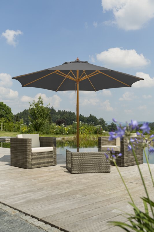 Schneider Malaga parasol in anthracite, 300 cm in diameter, on a wooden terrace with lounge furniture by a natural swimming pool. A blooming purple flower is in the foreground, with meadow, shrubs and forest in the background.