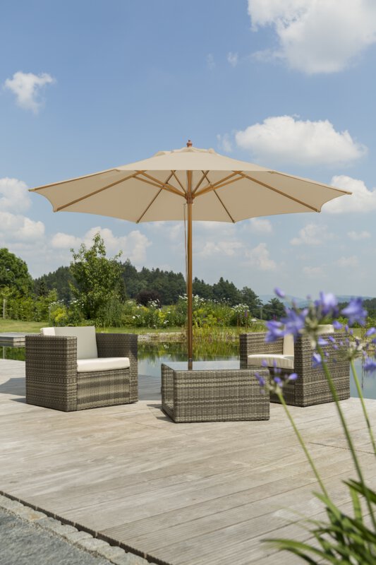 A Schneider Malaga parasol in ecru, 300 cm in diameter, sits on a wooden terrace with lounge furniture by a natural swimming pool. A blooming purple flower is in the foreground, with meadows, shrubs and a forest in the background.