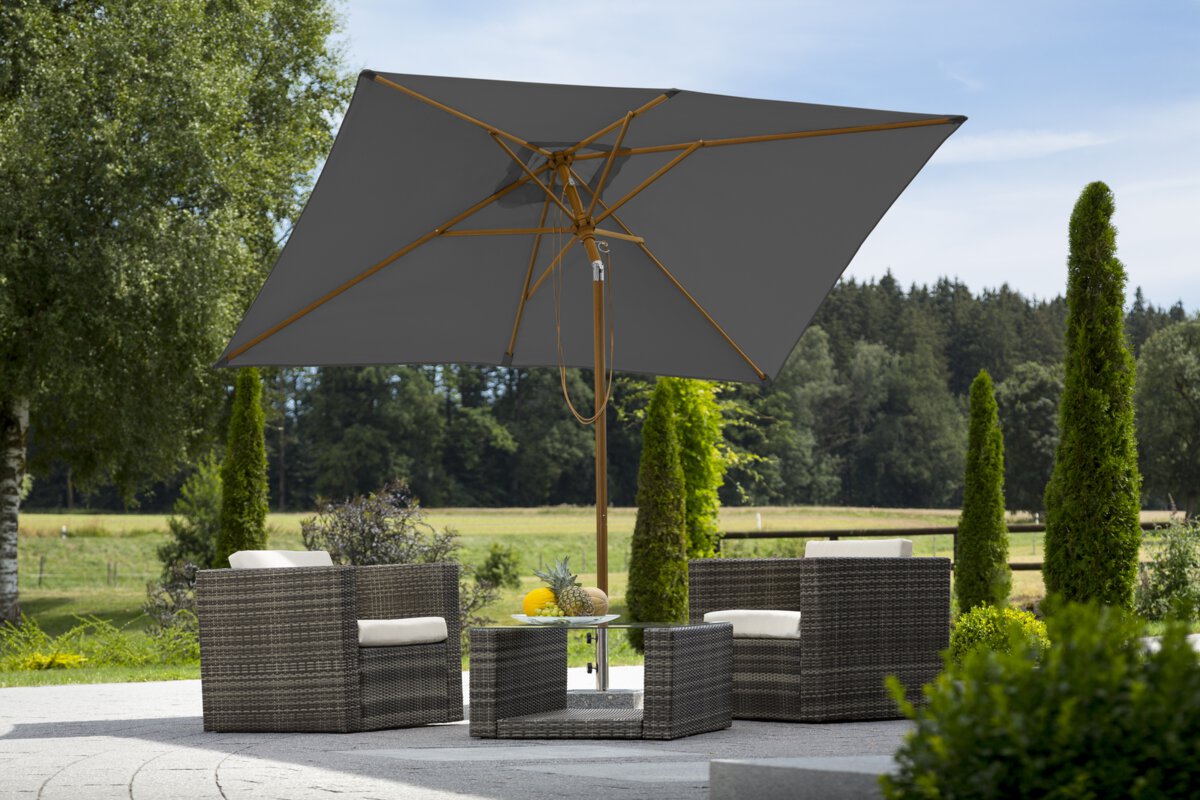 Schneider parasol Malaga in anthracite, 300x200cm, on a terrace with lounge furniture. A fruit bowl sits on the table. In the background are shrubs, a fence, a meadow with trees and a forest.