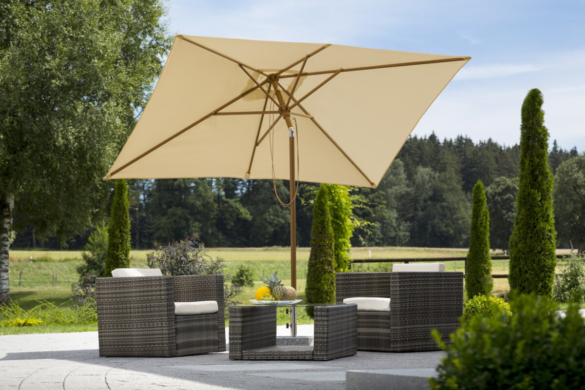 Schneider Malaga parasol in ecru, 300x200cm, on a terrace with lounge furniture. A fruit bowl sits on the table. In the background are shrubs, a fence, a meadow with trees and a forest.