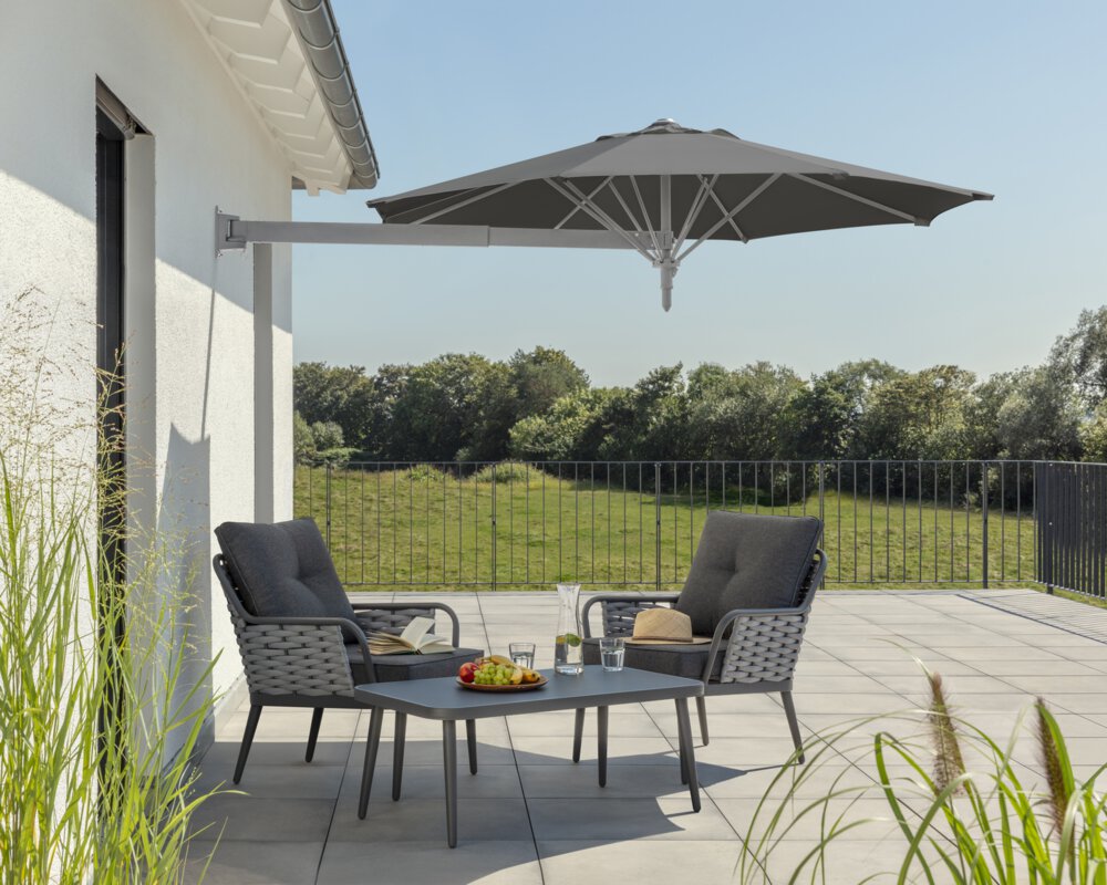 Schneider Sonnenschirm Muro in Anthrazit, 250 cm Durchmesser, auf einer Dachterrasse. Im Schatten des Schirms stehen zwei Stühle mit grauen Sitzpolstern und ein kleiner Tisch mit Obstschale, Karaffe und Gläsern. Auf den Stühlen ein aufgeschlagenes Buch und ein Hut. Im Hintergrund Wiese und Wald unter strahlend blauem Himmel.