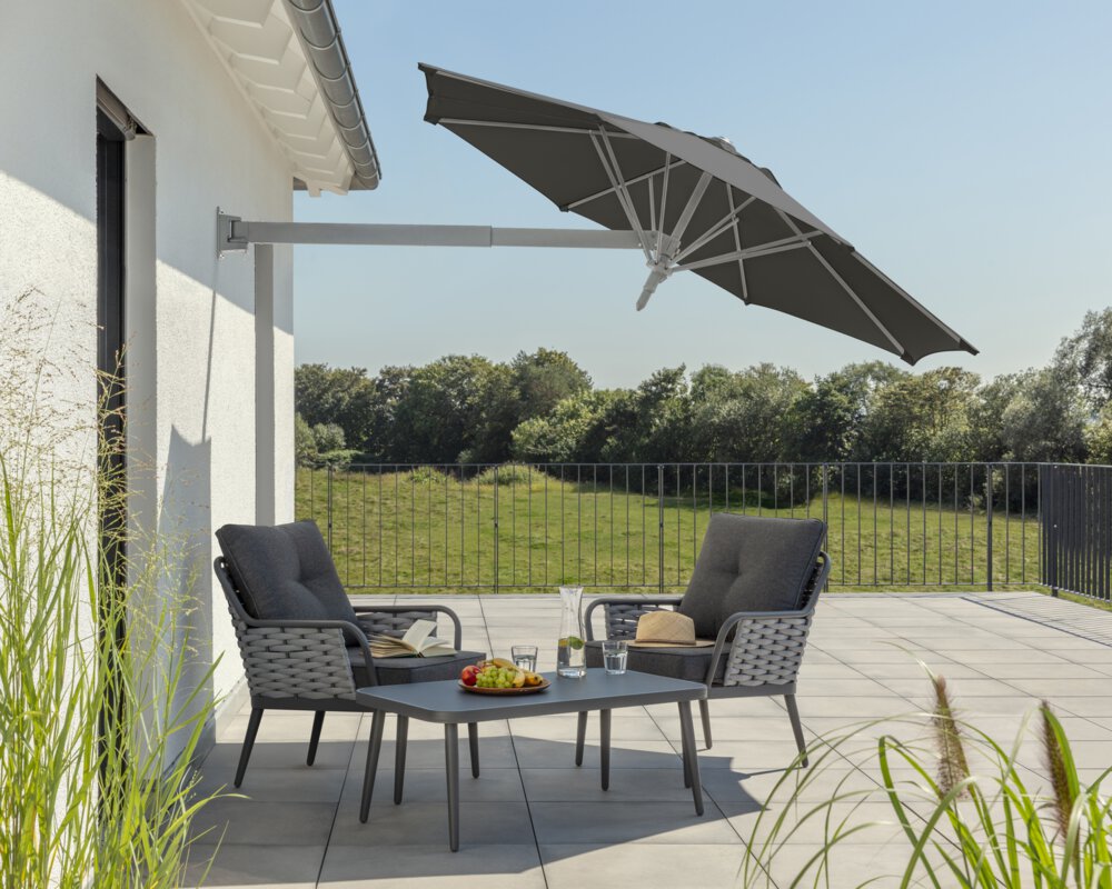 Schneider Sonnenschirm Muro in Anthrazit, 250 cm Durchmesser, geneigt, auf einer Dachterrasse. Im Schatten des Schirms stehen zwei Stühle mit grauen Sitzpolstern und ein kleiner Tisch mit Obstschale, Karaffe und Gläsern. Auf den Stühlen ein aufgeschlagenes Buch und ein Hut. Im Hintergrund Wiese und Wald unter strahlend blauem Himmel.