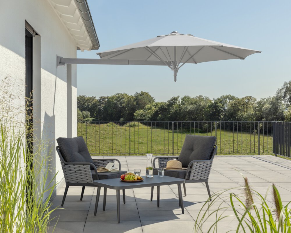 Schneider Sonnenschirm Muro in Silbergrau, 250 cm Durchmesser, auf einer Dachterrasse. Im Schatten des Schirms stehen zwei Stühle mit grauen Sitzpolstern und ein kleiner Tisch mit Obstschale, Karaffe und Gläsern. Auf den Stühlen ein aufgeschlagenes Buch und ein Hut. Im Hintergrund Wiese und Wald unter strahlend blauem Himmel.