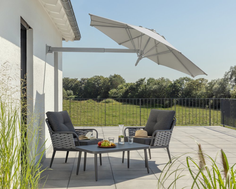 Schneider Sonnenschirm Muro in Silbergrau, 250 cm Durchmesser, geneigt, auf einer Dachterrasse. Im Schatten des Schirms stehen zwei Stühle mit grauen Sitzpolstern und ein kleiner Tisch mit Obstschale, Karaffe und Gläsern. Auf den Stühlen ein aufgeschlagenes Buch und ein Hut. Im Hintergrund Wiese und Wald unter strahlend blauem Himmel.