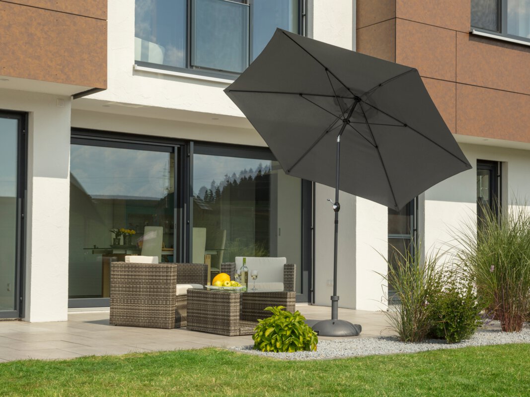 Schneider Napoli parasol in anthracite, 270 cm in diameter, on a terrace with lounge furniture. On the table is a fruit bowl with a bottle and glasses. Small shrubs and a meadow are in the foreground.