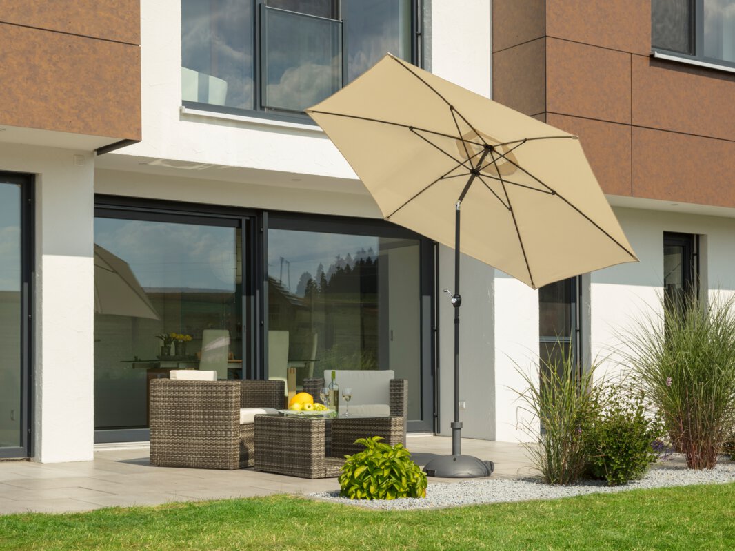 A Schneider Napoli parasol in ecru, 270 cm in diameter, sits on a terrace with lounge furniture. A fruit bowl with a bottle of wine and wine glasses sits on the table. Small shrubs and a meadow are in the foreground.
