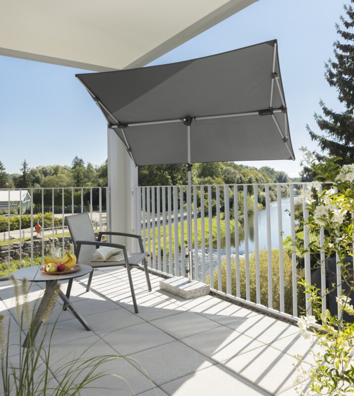 Schneider Novara parasol in anthracite, 190x140 cm, on a balcony with a metal railing. A chair and a small, round table with a fruit plate stand in the shade of the parasol. An open book lies on the chair. In the background, a river lined with numerous trees.
