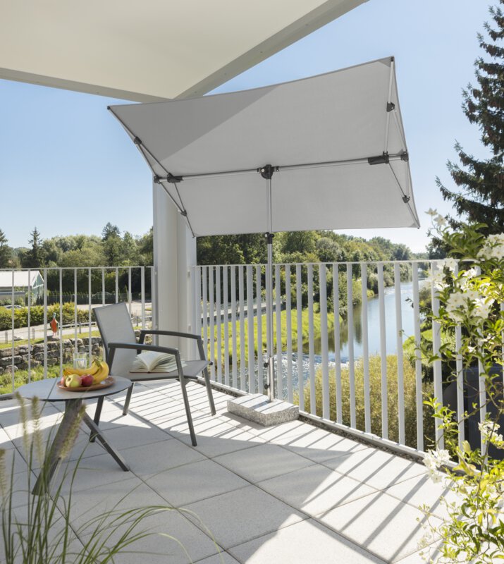 Schneider Novara parasol in silver grey, 190x140 cm, on a balcony with a metal railing. A chair and a small, round table with a fruit plate stand in the shade of the parasol. An open book lies on the chair. In the background, a river lined with numerous trees.