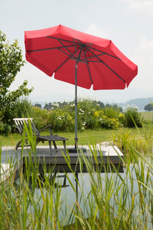 A red Schneider Orlando parasol, 270 cm in diameter, placed on a wooden deck by a natural swimming pool. A deck chair sits beneath it. Reeds are in the foreground and meadows and shrubs are in the background.