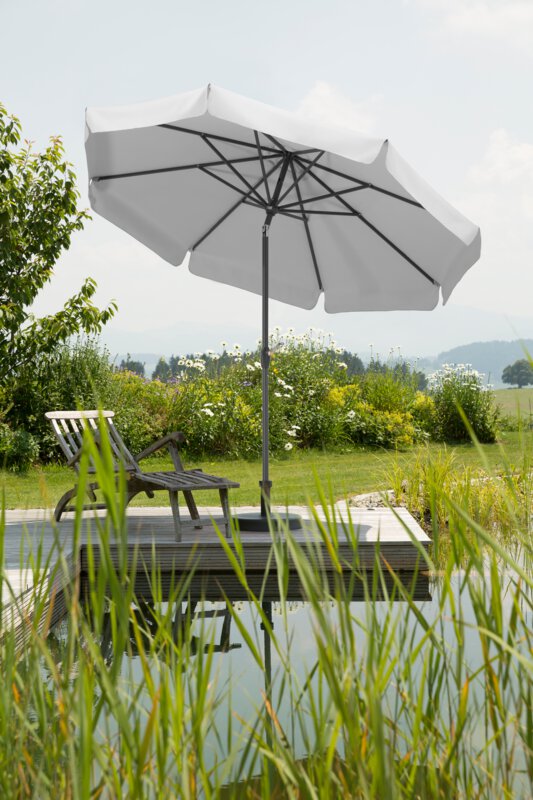 A Schneider Orlando parasol in silver grey, 270 cm in diameter, placed on a wooden deck by a natural swimming pool. A deck chair sits beneath it. Reeds are in the foreground and meadows and shrubs are in the background.