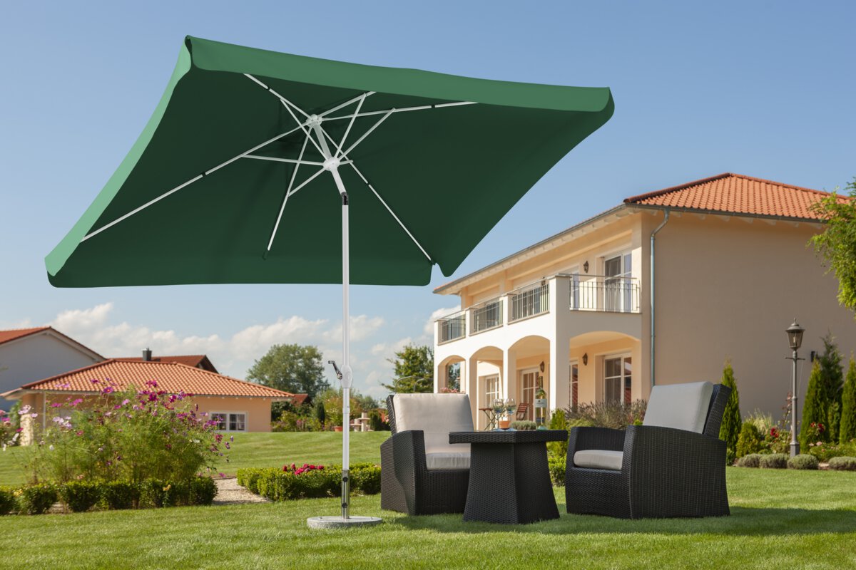 Schneider Oslo parasol in green, 200x300 cm, on a lawn above lounge furniture. In the background blooming flowers, shrubs and houses in Mediterranean style.