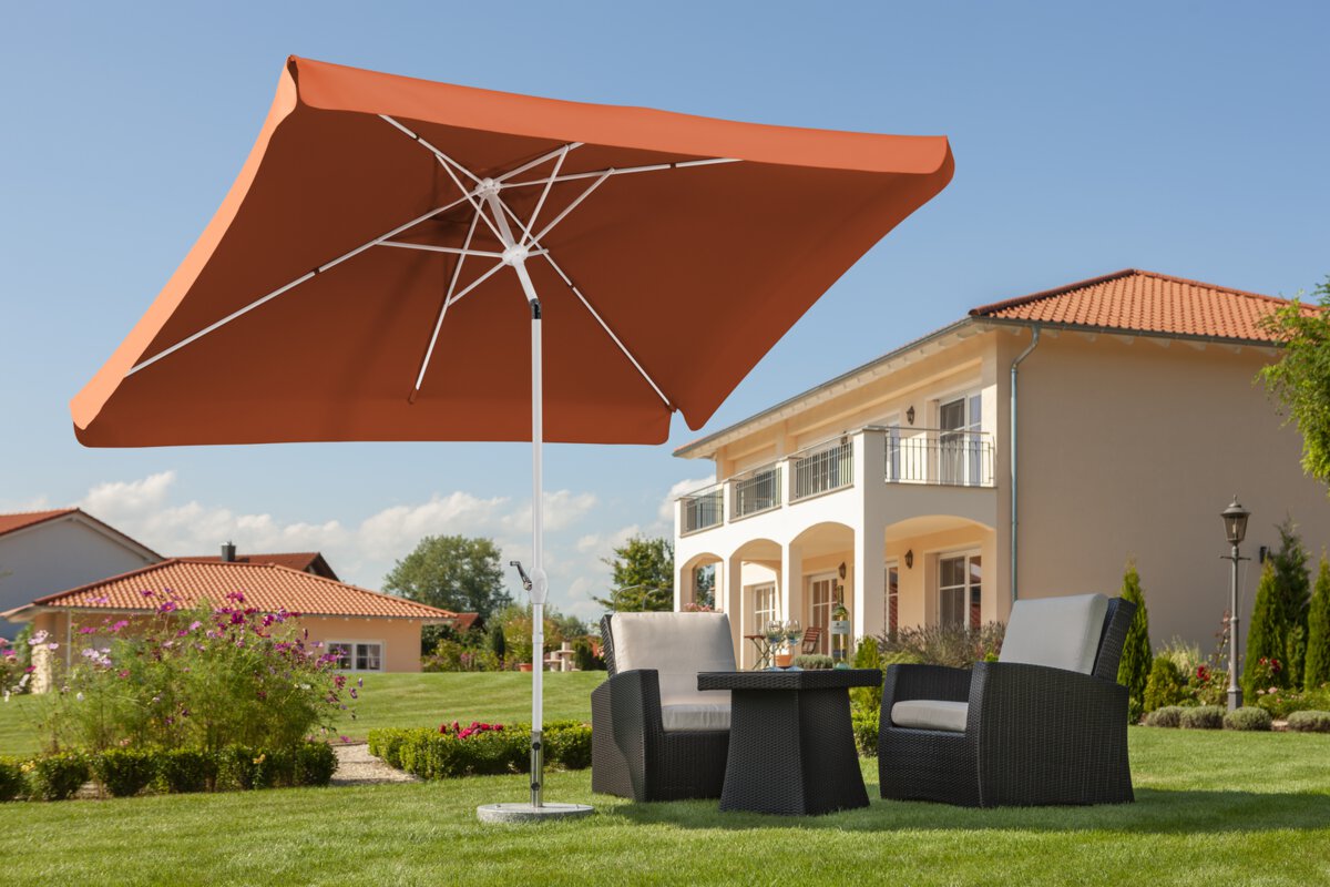 Schneider Oslo parasol in terracotta, 200x300 cm, on a lawn above lounge furniture. In the background blooming flowers, shrubs and houses in Mediterranean style.