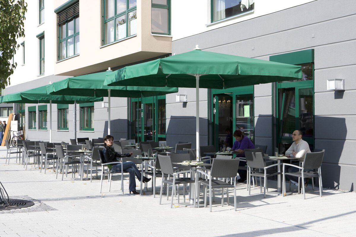 Vier Schneider Sonnenschirme Quadro in Grün, 300x300 cm vor einem Restaurant. Darunter stehen Tische und Stühle, an denen vereinzelt Gäste Platz genommen haben.