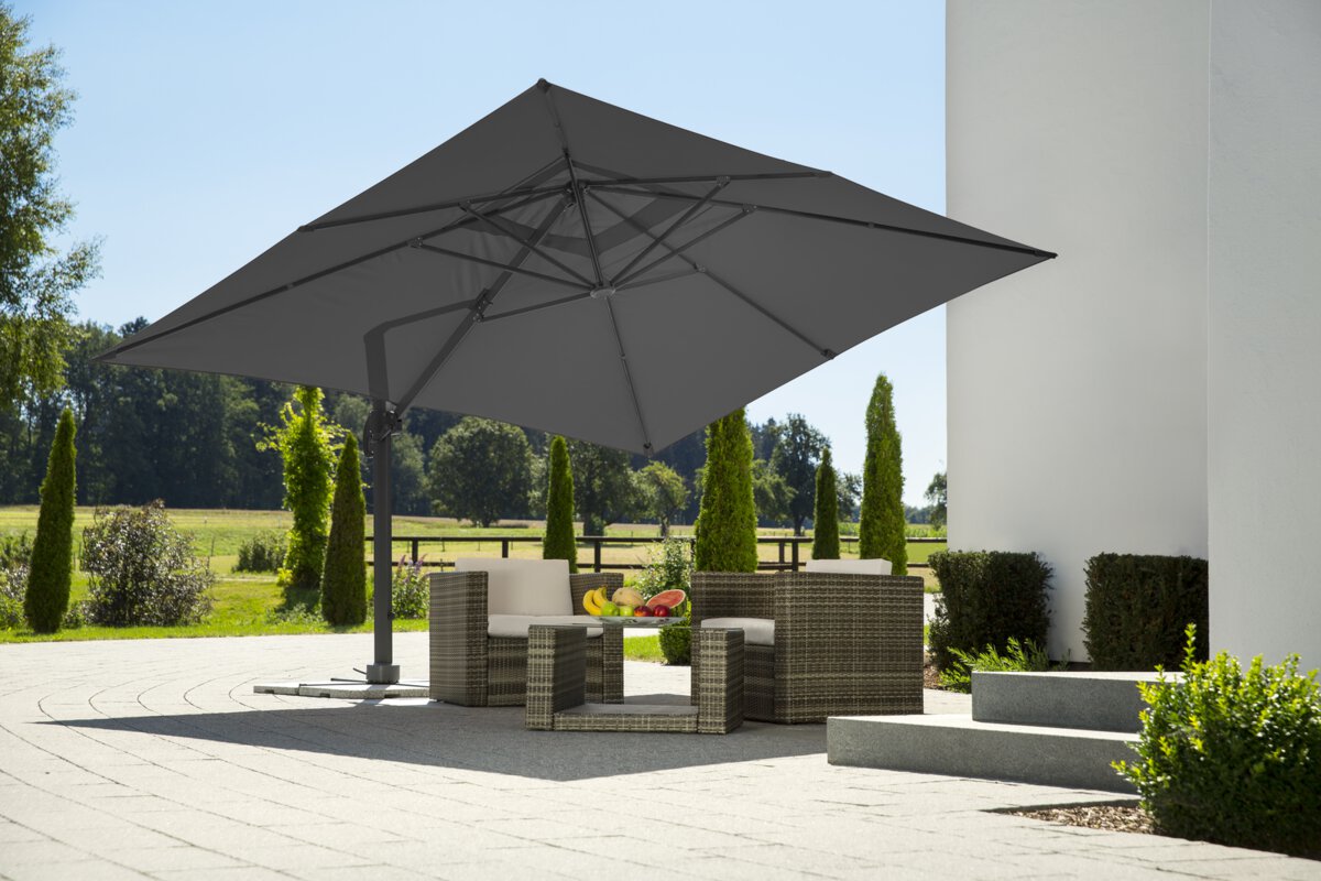 Schneider Rhodos Grande parasol in anthracite, 400x300 cm in diameter, on a terrace with lounge furniture. A fruit bowl sits on the table. In the background, shrubs, a fence, a meadow with trees and a forest.