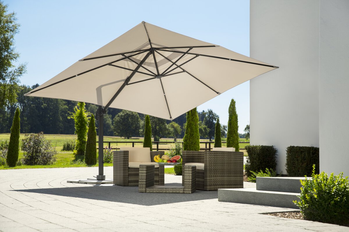 Schneider Rhodos Grande parasol in ecru, 400x300 cm in diameter, on a terrace with lounge furniture. A fruit bowl sits on the table. In the background are shrubs, a fence, a meadow with trees, and a forest.