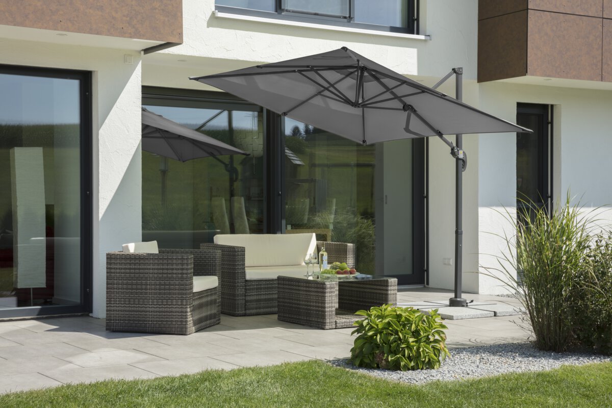 Schneider parasol Rhodos Junior in anthracite, 230x230 cm, inclined, on a terrace with lounge furniture. A fruit bowl, a bottle and glasses are on the table. Small shrubs and a meadow are in the foreground.