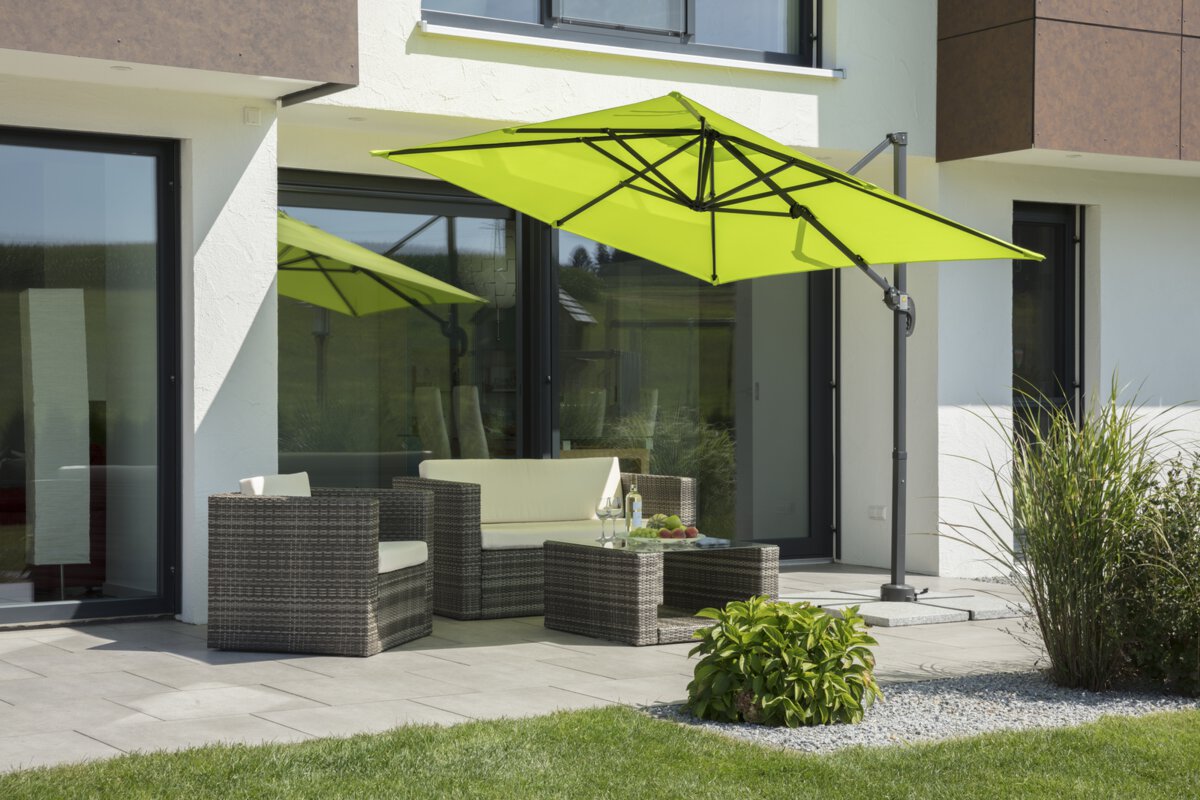 Schneider Rhodos Junior parasol in apple green, 230x230 cm, inclined, on a terrace with lounge furniture. A fruit bowl, a bottle, and glasses are on the table. Small shrubs and a meadow are in the foreground.