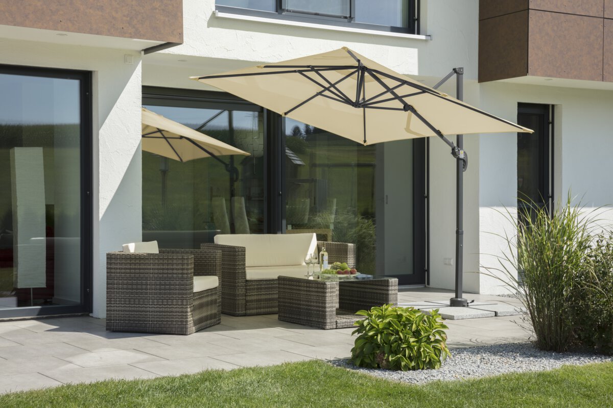 Schneider parasol Rhodos Junior in ecru, 230x230 cm, inclined, on a terrace with lounge furniture. A fruit bowl, a bottle, and glasses are on the table. Small shrubs and a meadow are in the foreground.
