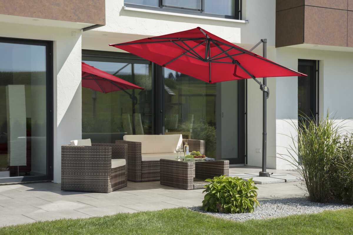 Schneider parasol Rhodos Junior in red, 230x230 cm, inclined, on a terrace with lounge furniture. A fruit bowl, a bottle and glasses are on the table. Small shrubs and a meadow are in the foreground.