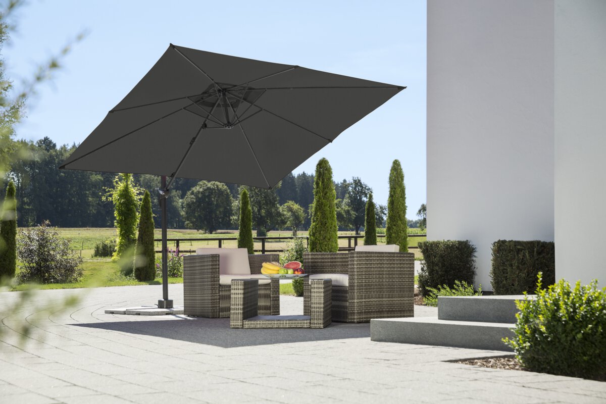 Schneider parasol Rhodos Junior in anthracite, 270x270 cm, on a terrace with lounge furniture. A fruit bowl sits on the table. In the background are shrubs, a fence, a meadow with trees and a forest.