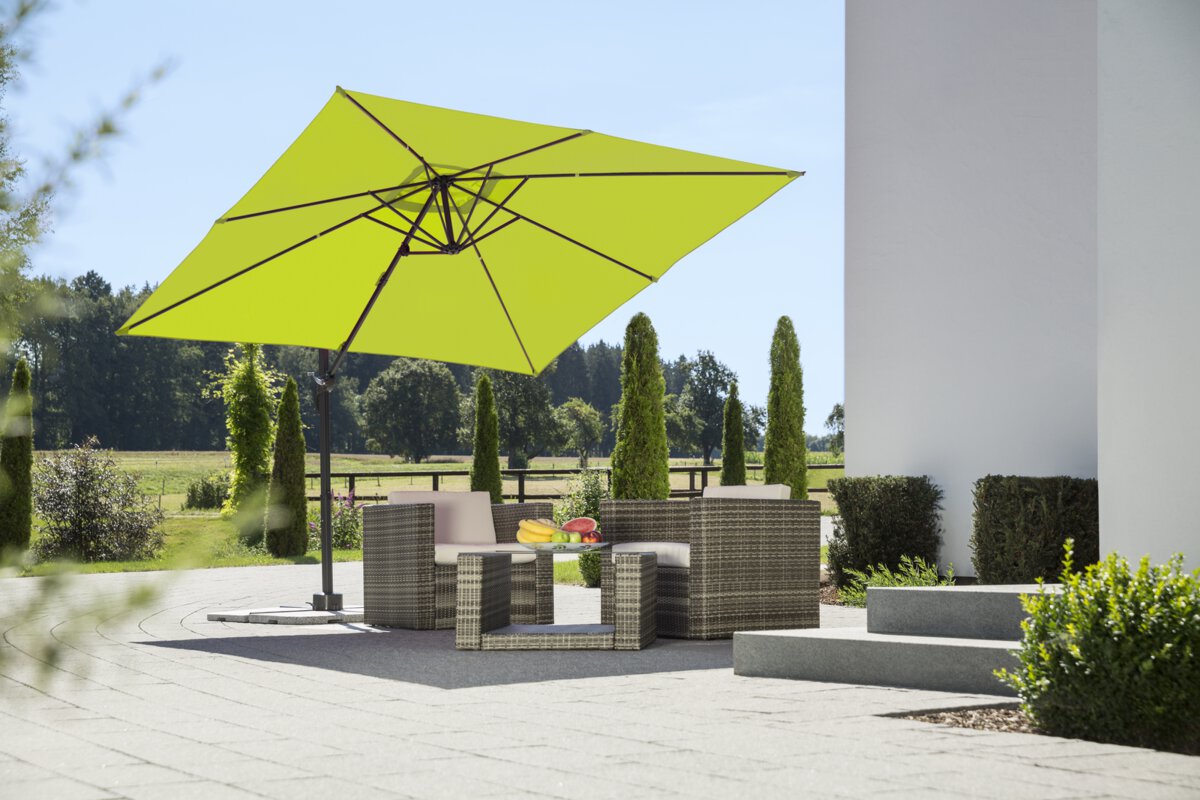 Schneider Rhodos Junior parasol in apple green, 270x270 cm, on a terrace with lounge furniture. A fruit bowl sits on the table. In the background are shrubs, a fence, a meadow with trees and a forest.
