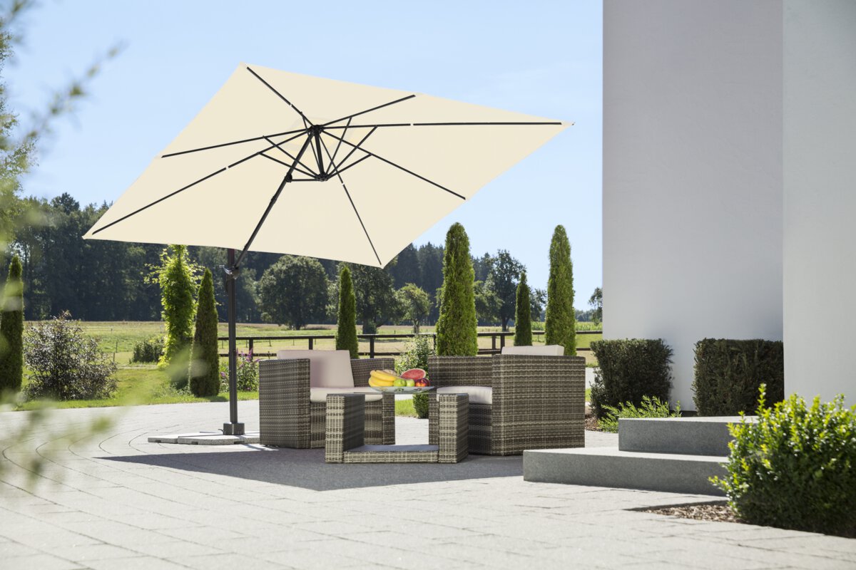 Schneider parasol Rhodos Junior in ecru, 270x270 cm, on a terrace with lounge furniture. A fruit bowl sits on the table. In the background are shrubs, a fence, a meadow with trees and a forest.
